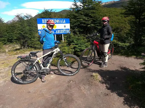 Dos riders con casco y sus e-Bikes junto al cartel de bienvenida a la República Argentina, en el bosque